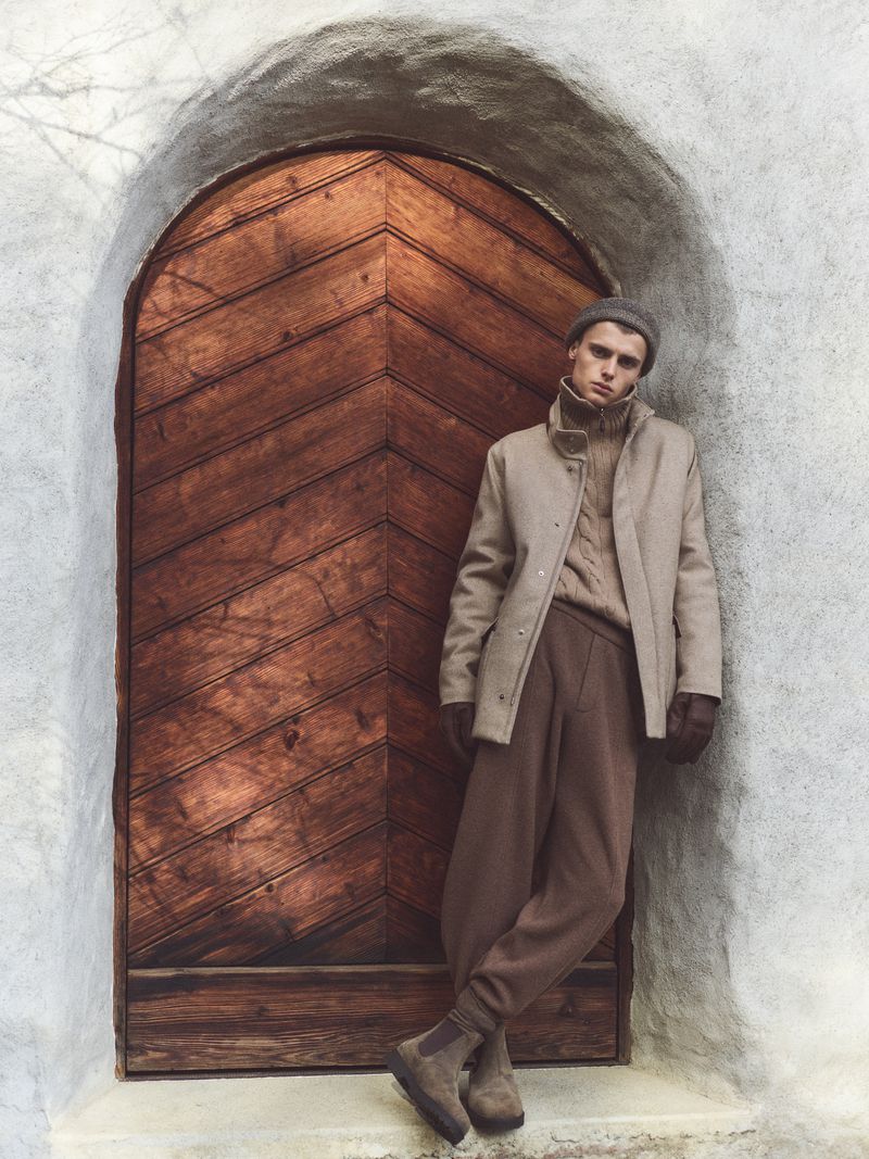 Male fashion model poses in front of an arched wooden door in a textured beige coat, brown sweater, trousers, beanie, and suede boots.