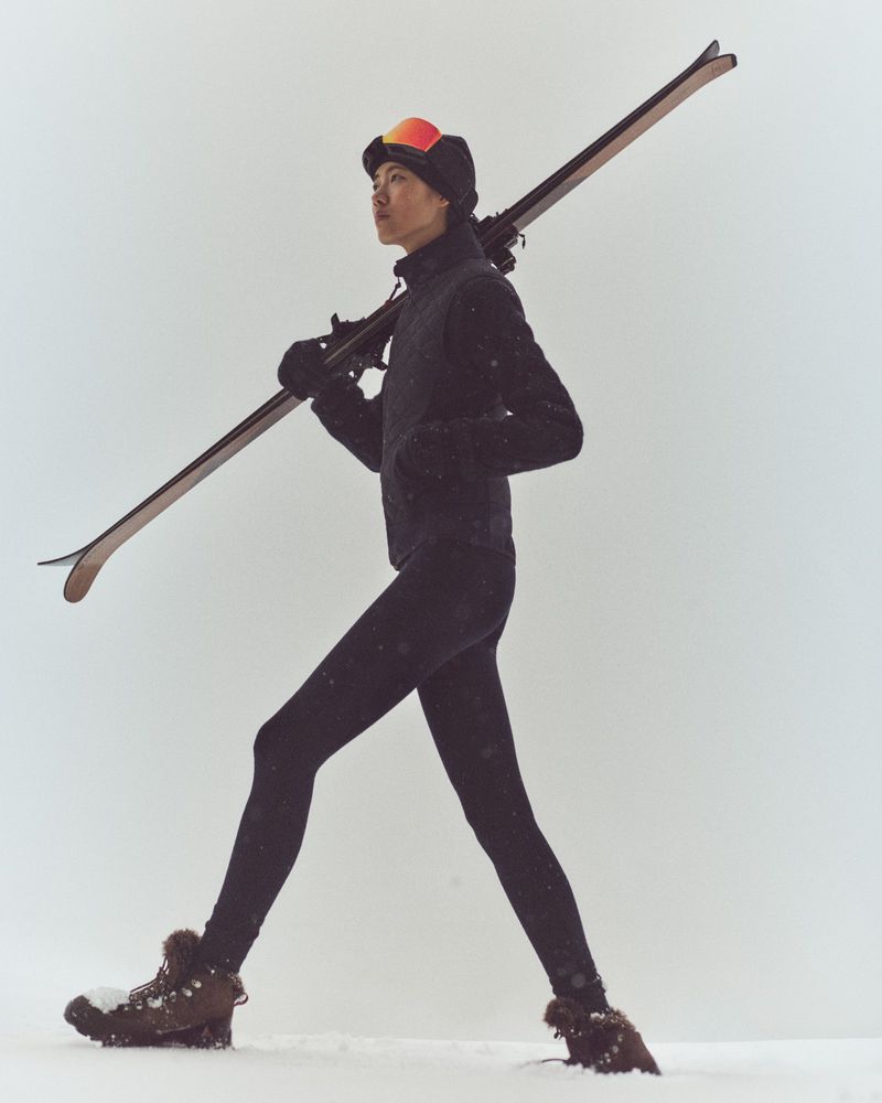 Athletic woman in winter clothing carrying skis over her shoulder walks through the snow on a cloudy day, looking ahead.