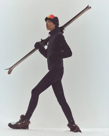 Athletic woman in winter clothing carrying skis over her shoulder walks through the snow on a cloudy day, looking ahead.