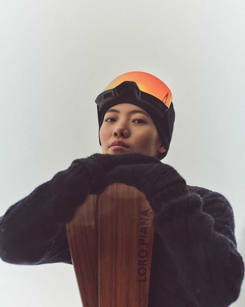 Portrait of an Asian female skier in a black beanie, goggles, and sweater, resting her arms on wood skis by Loro Piana.