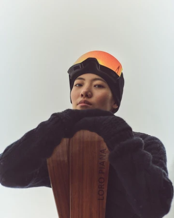 Portrait of an Asian female skier in a black beanie, goggles, and sweater, resting her arms on wood skis by Loro Piana.