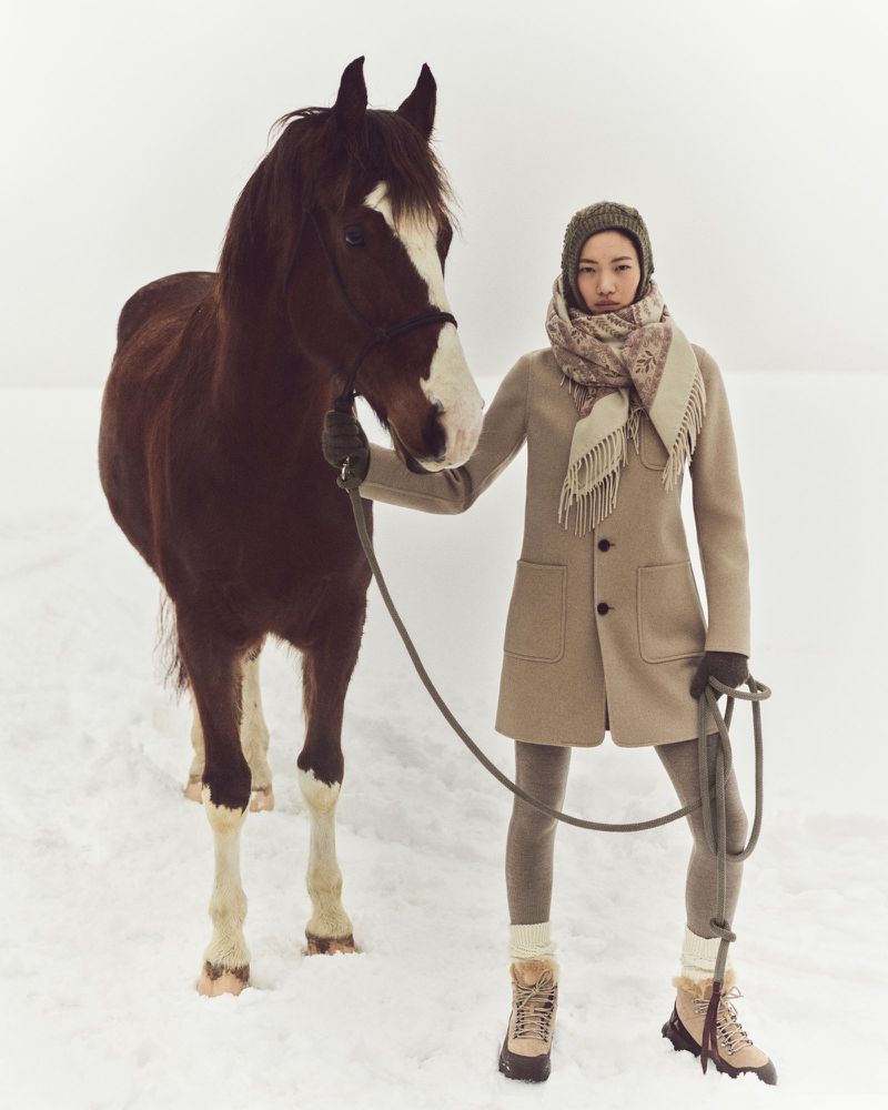 Woman in winter fashion posing with a brown horse in a snow-covered field, wearing a tan coat, scarf, hat, leggings, and hiking boots.