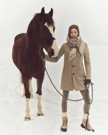 Woman in winter fashion posing with a brown horse in a snow-covered field, wearing a tan coat, scarf, hat, leggings, and hiking boots.
