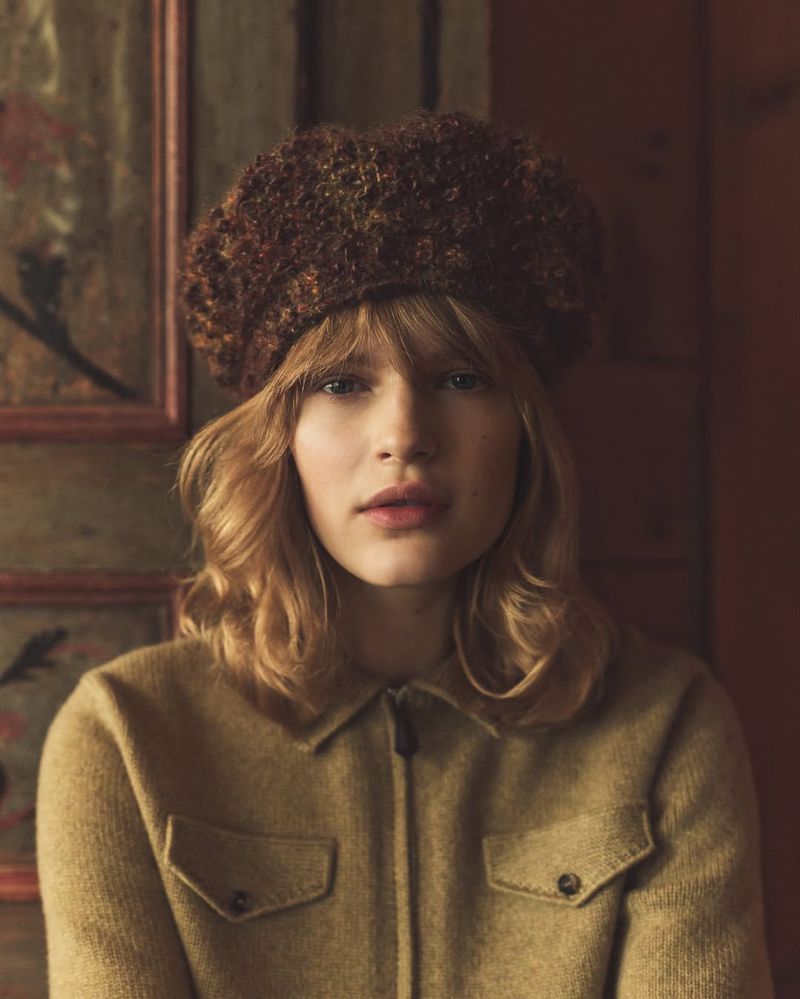 Portrait of a young woman wearing a textured brown hat and beige shirt against a softly painted, rustic backdrop in muted tones.