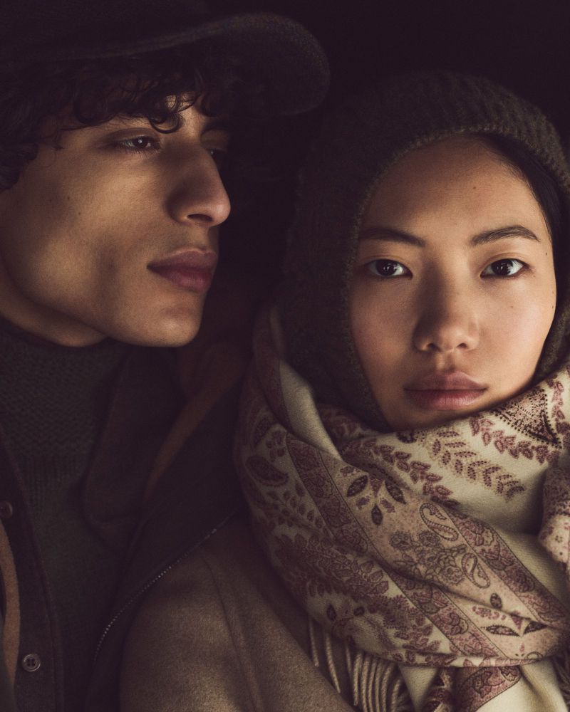 Portrait of a diverse couple in muted tones. The Asian woman, wrapped in a scarf, gazes directly at the camera. The man is looking off-frame.