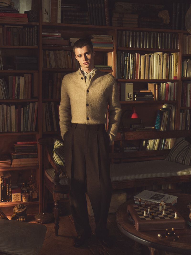 Young man in sweater and trousers stands indoors near bookshelf with chess set on table in warm lighting, evoking a scholarly atmosphere.