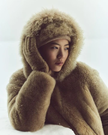 Model wearing a fuzzy hooded coat and matching hat against a snowy white background for Loro Piana Holiday 2025 Campaign