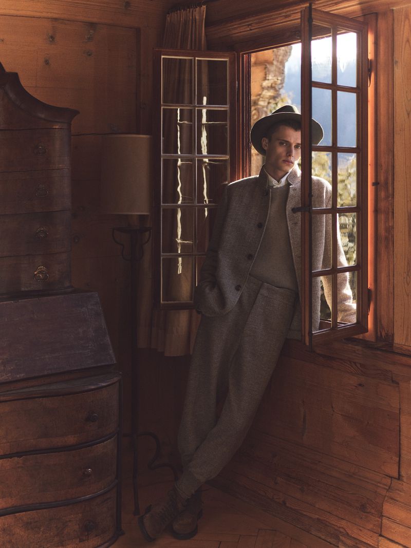 Man in tweed suit and hat leans in wooden window frame with mountain view, standing indoors, with dresser and lamp.