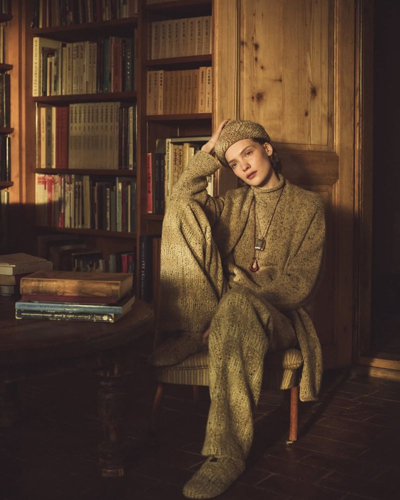 Woman in a matching knit sweater, beret, and trousers sits in front of a bookshelf in a room with dark wood floors and walls.