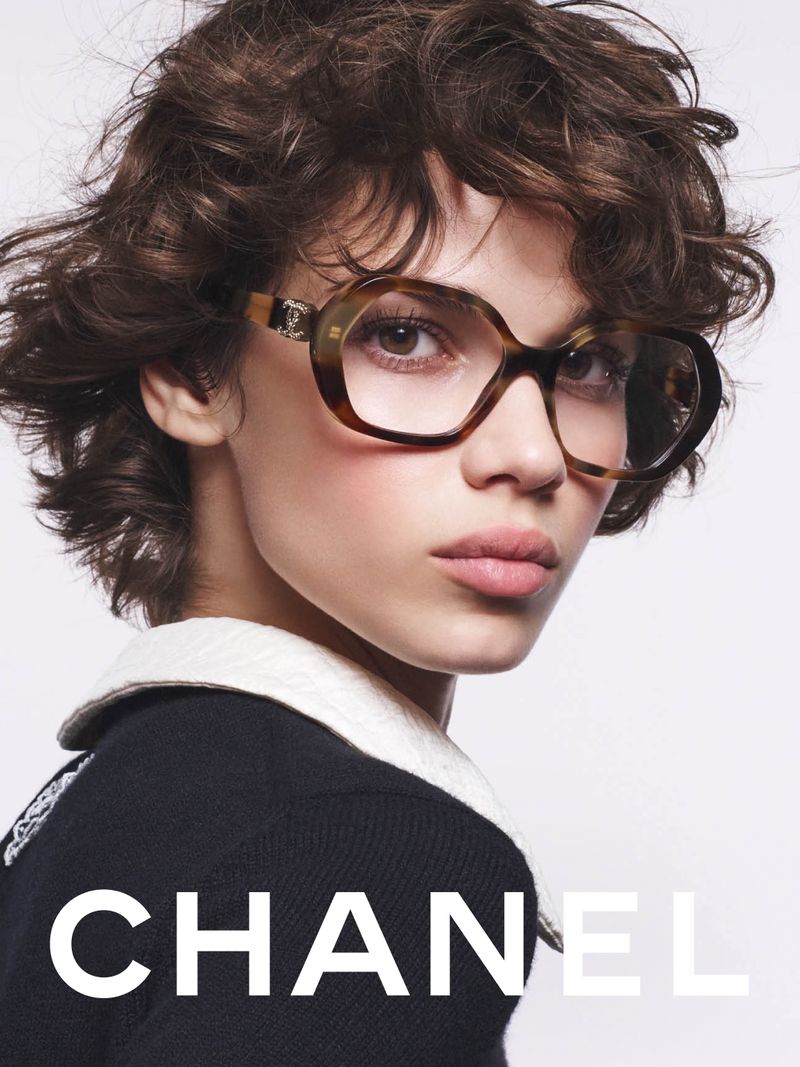 Close-up shot of a woman with brown curly hair wearing stylish brown-framed glasses and a black and white top in a studio setting.