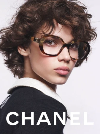 Close-up shot of a woman with brown curly hair wearing stylish brown-framed glasses and a black and white top in a studio setting.