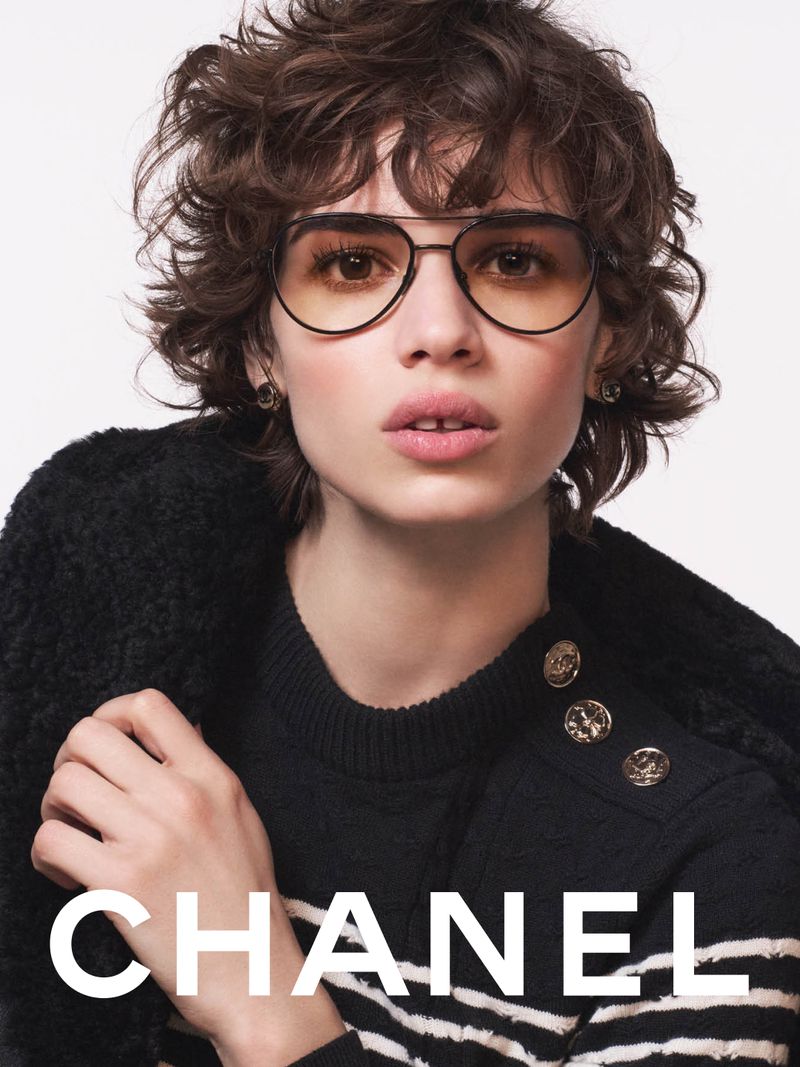 Woman in sunglasses, black wool jacket, and black and white striped shirt, posing in a fashion advertisement for Chanel.