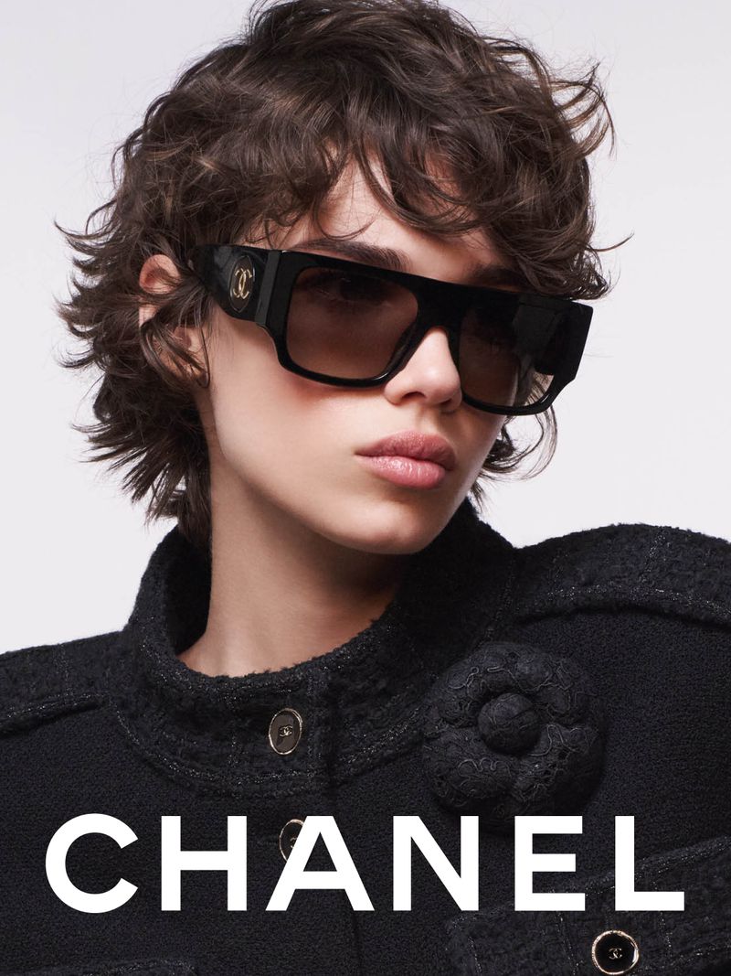 Fashion shot of a woman wearing oversized black sunglasses and a black jacket with a flower embellishment, with the CHANEL logo.