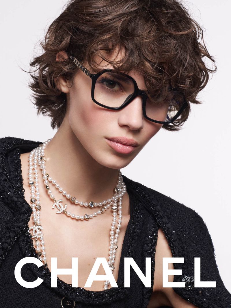 A woman with dark curly hair and glasses poses in a fashion shot, wearing a pearl necklace and a black textured jacket against a white background.