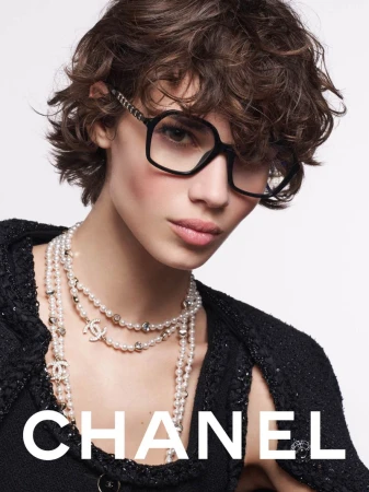 A woman with dark curly hair and glasses poses in a fashion shot, wearing a pearl necklace and a black textured jacket against a white background.