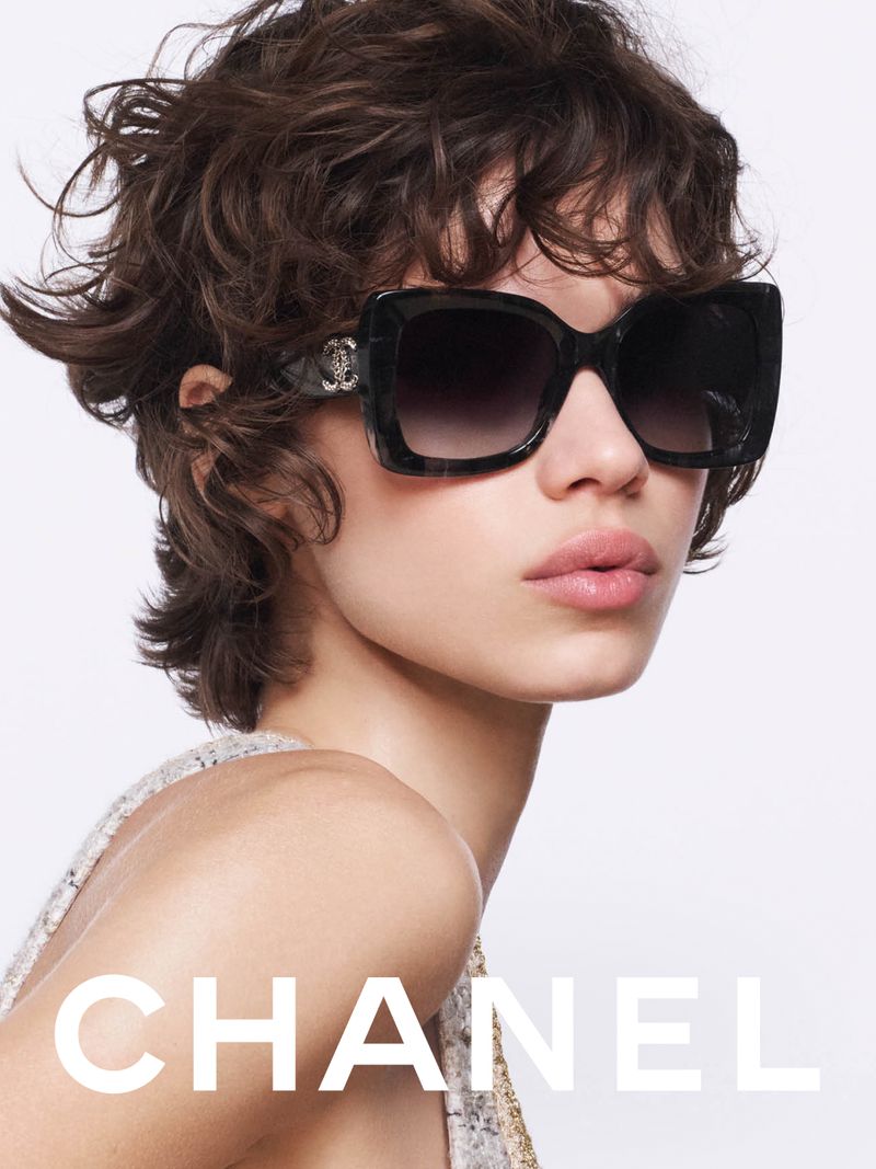 Close-up of a model wearing black, oversized sunglasses. The glasses have a light-colored trim with the CHANEL logo in silver. 