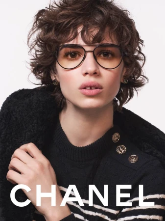 Model with short curly brown hair wearing aviator glasses and a dark sweater against a clean background