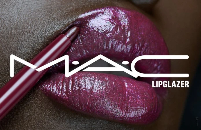 Close-up of an adult female with dark skin and purple lipstick, holding a matching lip pencil, featuring the MAC logo and 'Lipglazer' text.