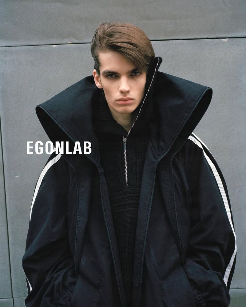Portrait of a young man wearing a black coat with large collar and white stripes, posing outdoors against a concrete wall.