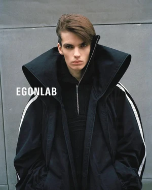 Young man wearing an oversized black coat with a wide collar and white stripe details against a grey concrete wall background.