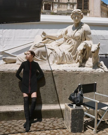 Fashion shot outdoors with a woman posing in a black dress and thigh-high boots next to a large neoclassical sculpture in daylight.