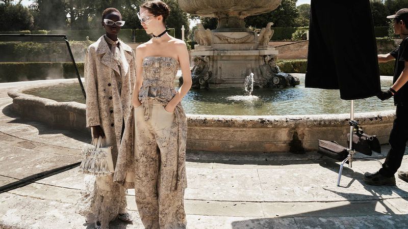 Two fashion models pose outdoors near a large stone fountain, showcasing patterned coats and dresses in a daytime editorial shoot.