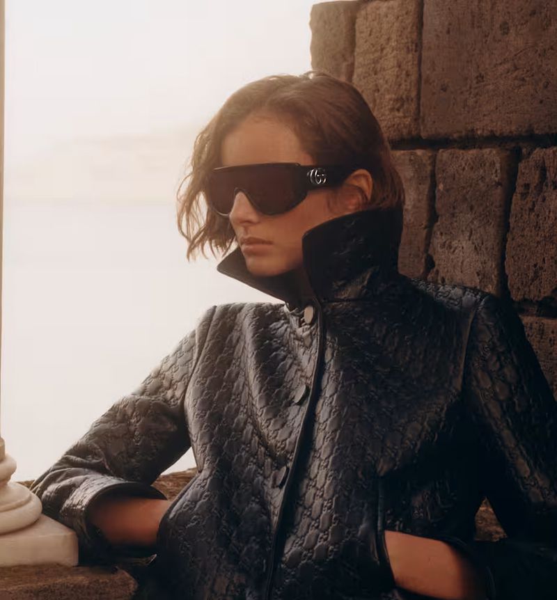 Fashion shot of a woman in sunglasses and a black patterned jacket posing next to a stone pillar in an outdoor setting.