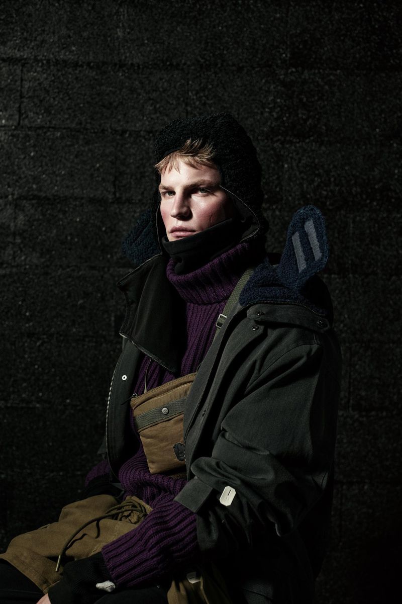 A fair-skinned male model poses in a purple turtleneck sweater, dark jacket, and trapper hat against a dark, textured background.