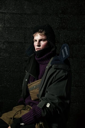 A fair-skinned male model poses in a purple turtleneck sweater, dark jacket, and trapper hat against a dark, textured background.