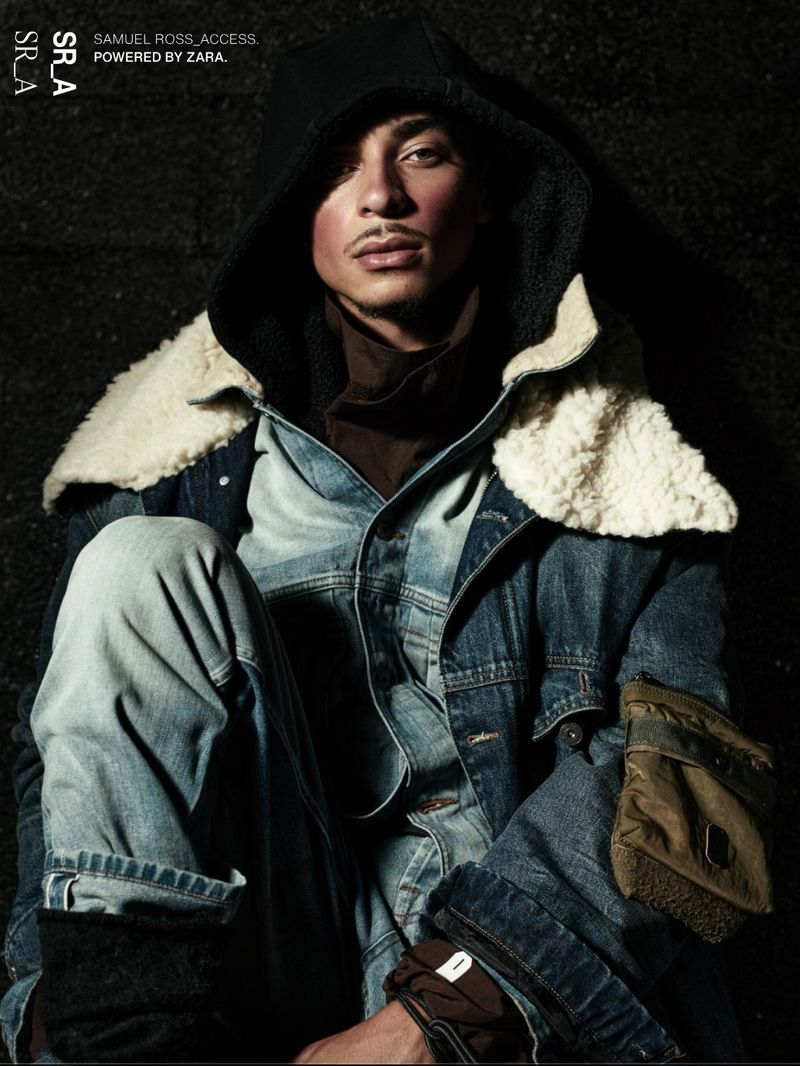 Man in denim jacket and jeans poses in dark studio, showcasing fashion and style with a blend of urban and contemporary aesthetics.