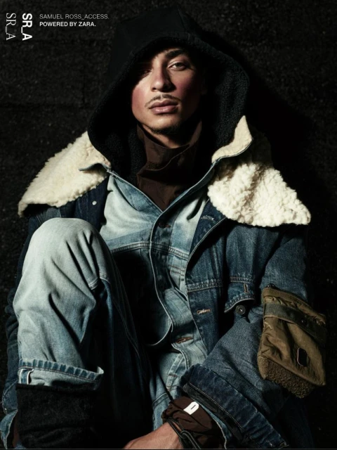 Man in denim jacket and jeans poses in dark studio, showcasing fashion and style with a blend of urban and contemporary aesthetics.