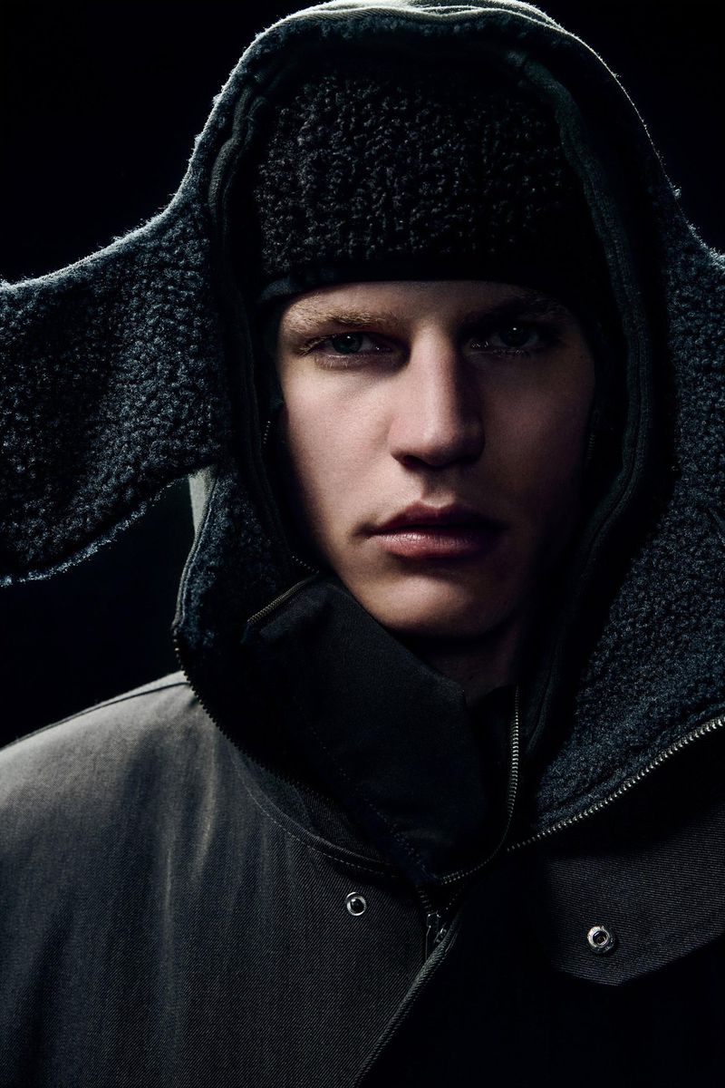 Close-up of a fair-skinned adult male wearing a dark hooded jacket and a black fur hat against a dark background in portrait orientation.