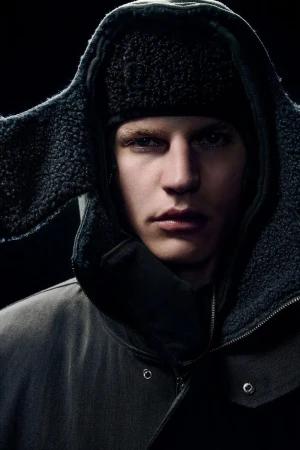 Close-up of a fair-skinned adult male wearing a dark hooded jacket and a black fur hat against a dark background in portrait orientation.