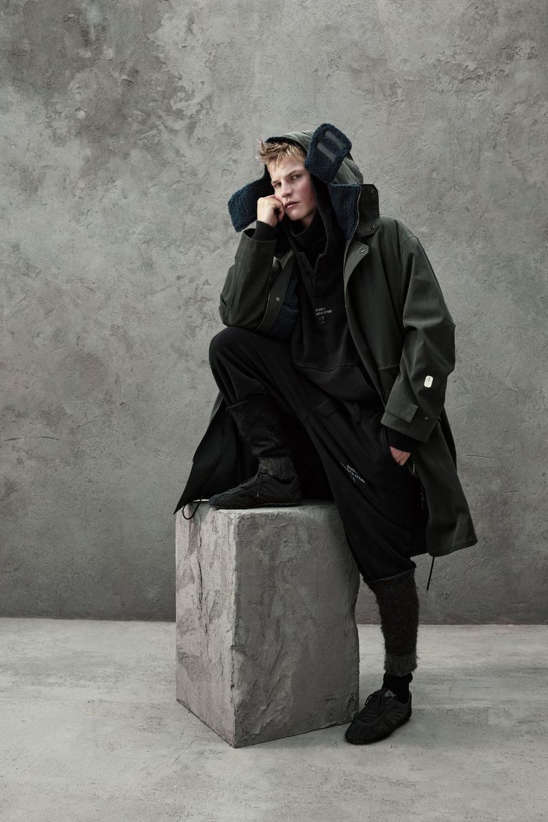 A blonde male model in a dark green hooded coat and black pants sits on a gray stone block against a mottled gray background in a studio shot.