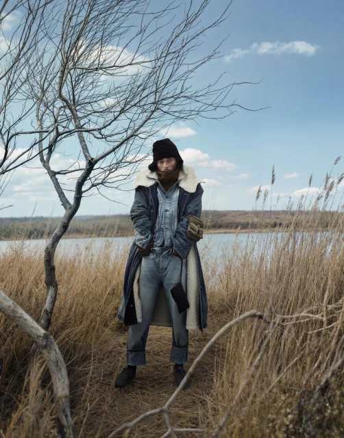 Man in denim and fur-lined coat standing in field of tall grass near lake under blue sky with clouds, framed by bare tree branches.