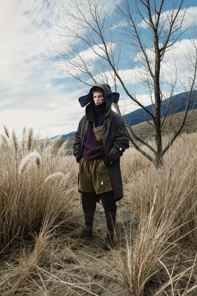 Model wearing a dark parka with ear-flap hood and purple sweater standing in tall dry grass in a meadow