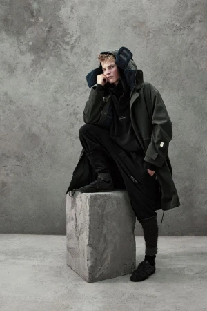 Model in dark green parka and black trousers squatting on a stone block against a textured concrete wall.