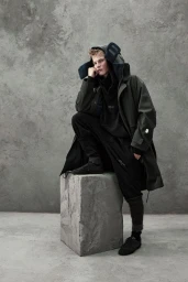 Model in dark green parka and black trousers squatting on a stone block against a textured concrete wall.