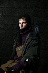 Model wearing a purple turtleneck, dark jacket, and hood against a dark textured block wall background