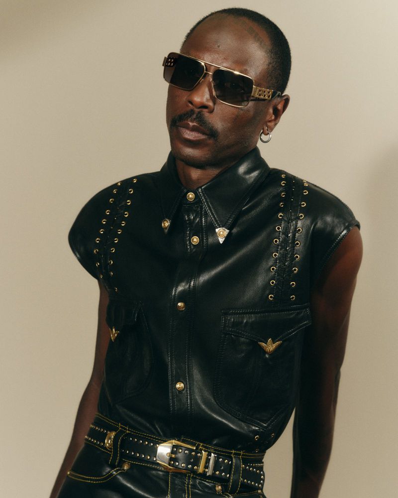 Black man in leather vest and sunglasses poses indoors. Head and shoulders shot, looking at the camera with neutral expression. 
