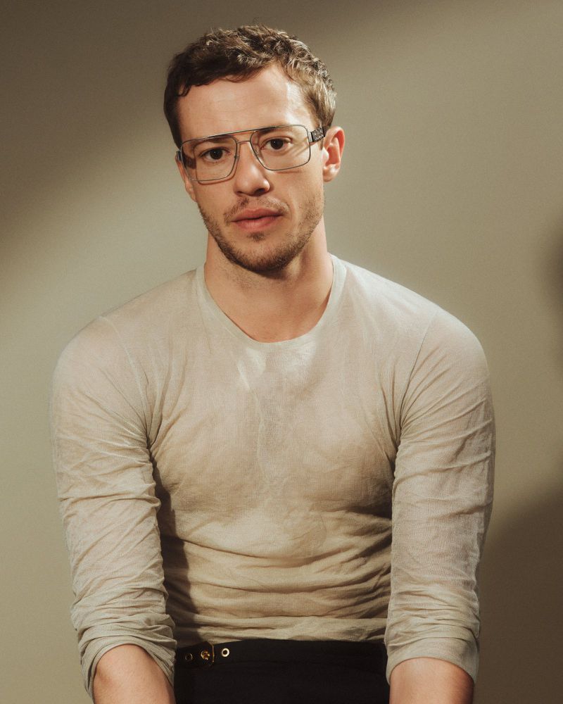 Man wearing wire-rimmed eyeglasses and a neutral long-sleeve top against a grey studio backdrop.