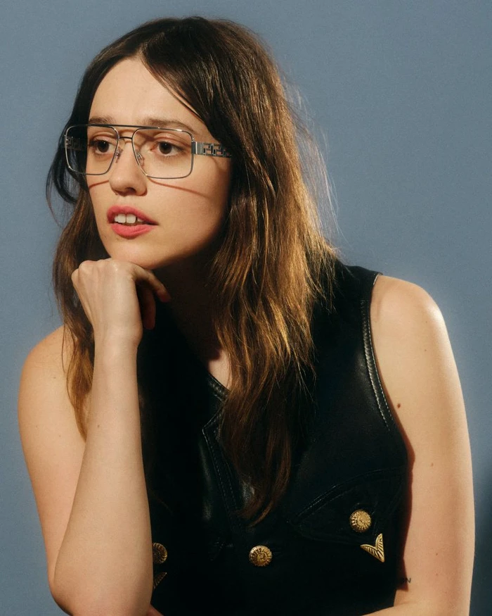 Aimee Lou Wood wearing metal aviator eyeglasses and a dark vest against a solid grey studio background.