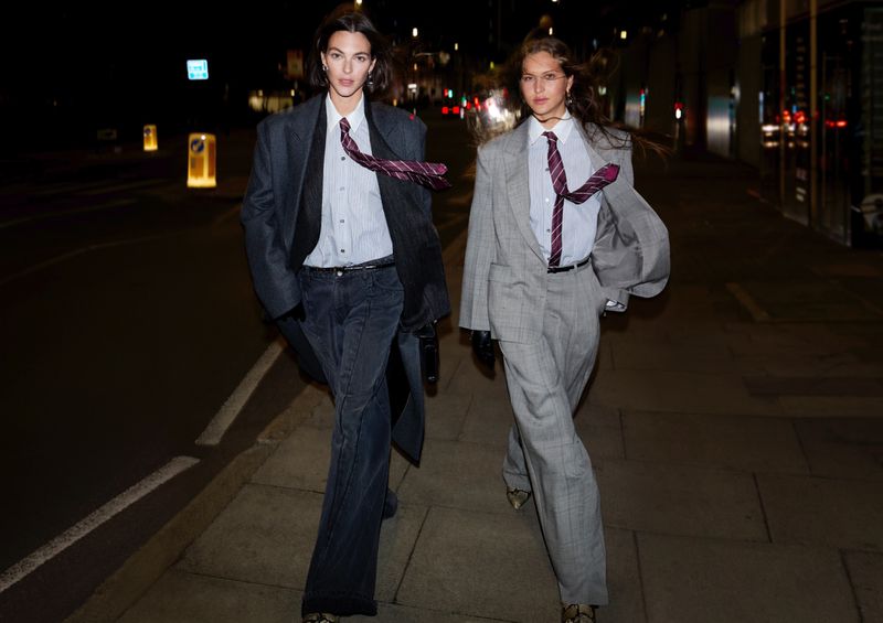 Two women in suits walk on a city sidewalk at night, wearing ties and fashionable outfits, under the dark sky with street lights.
