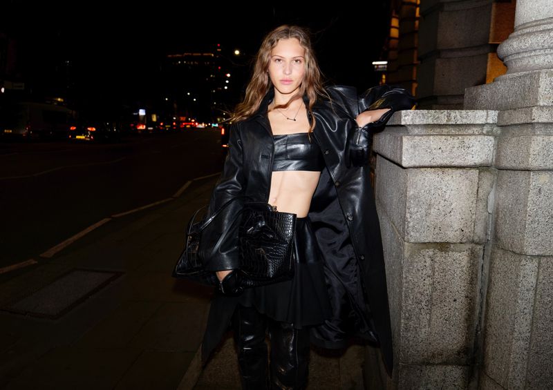 Woman in black leather top and coat poses at night in urban setting with dark background and city lights, holding a black handbag.