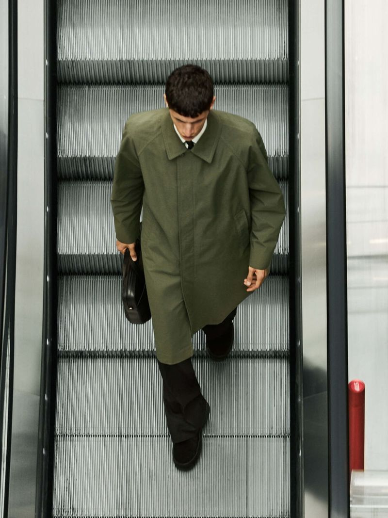 Overhead shot of an adult man on an escalator wearing a trench coat and holding a briefcase, walking down towards the camera.