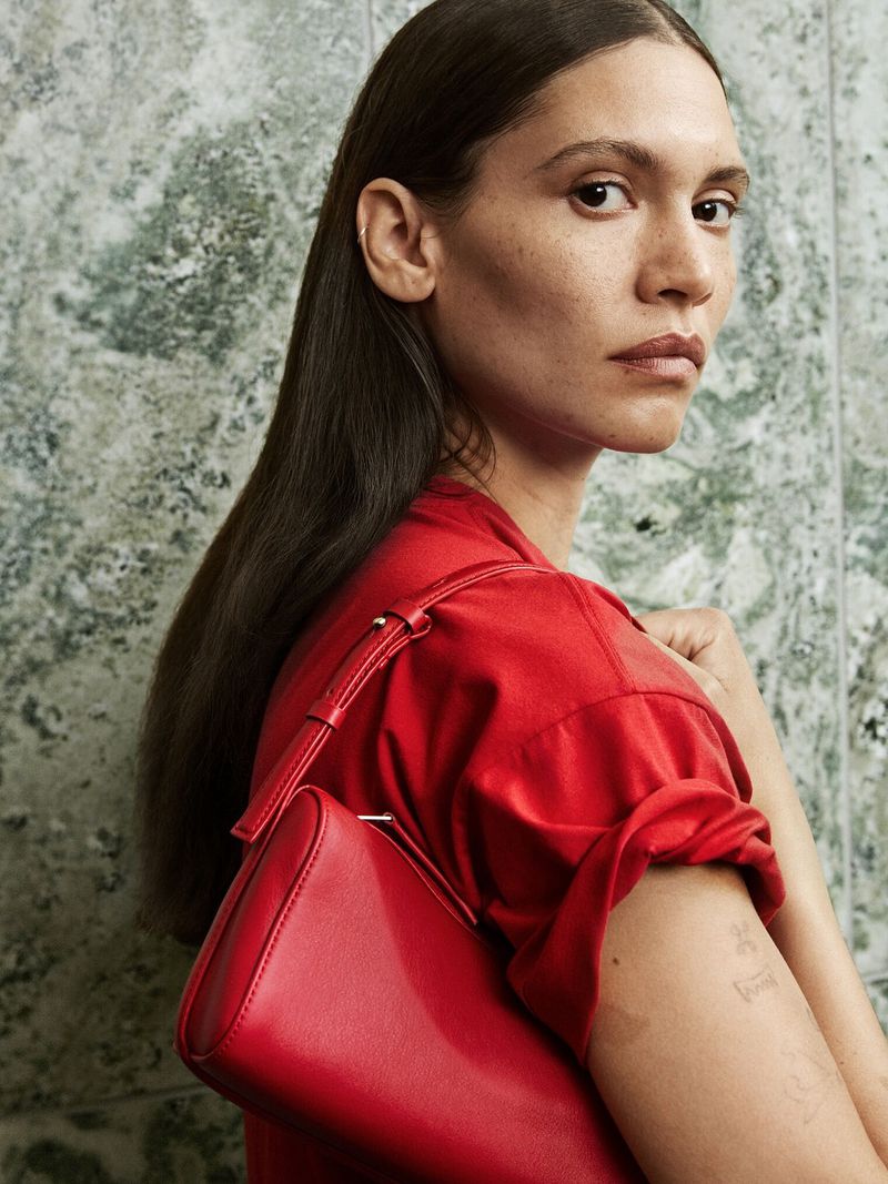 Brunette woman with a red handbag and matching short-sleeve shirt poses against a marble wall in a fashion shot.