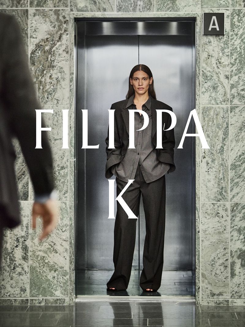 Fashion shot of a woman in a grey suit standing in an elevator with marble walls, a man's arm is partially visible in the foreground.