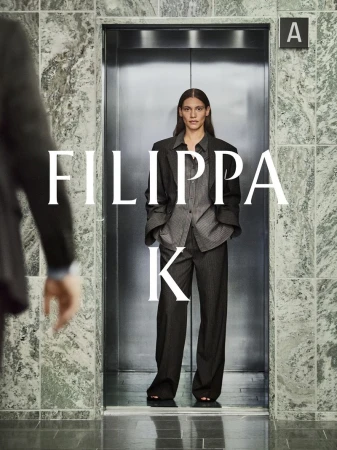 Fashion shot of a woman in a grey suit standing in an elevator with marble walls, a man's arm is partially visible in the foreground.