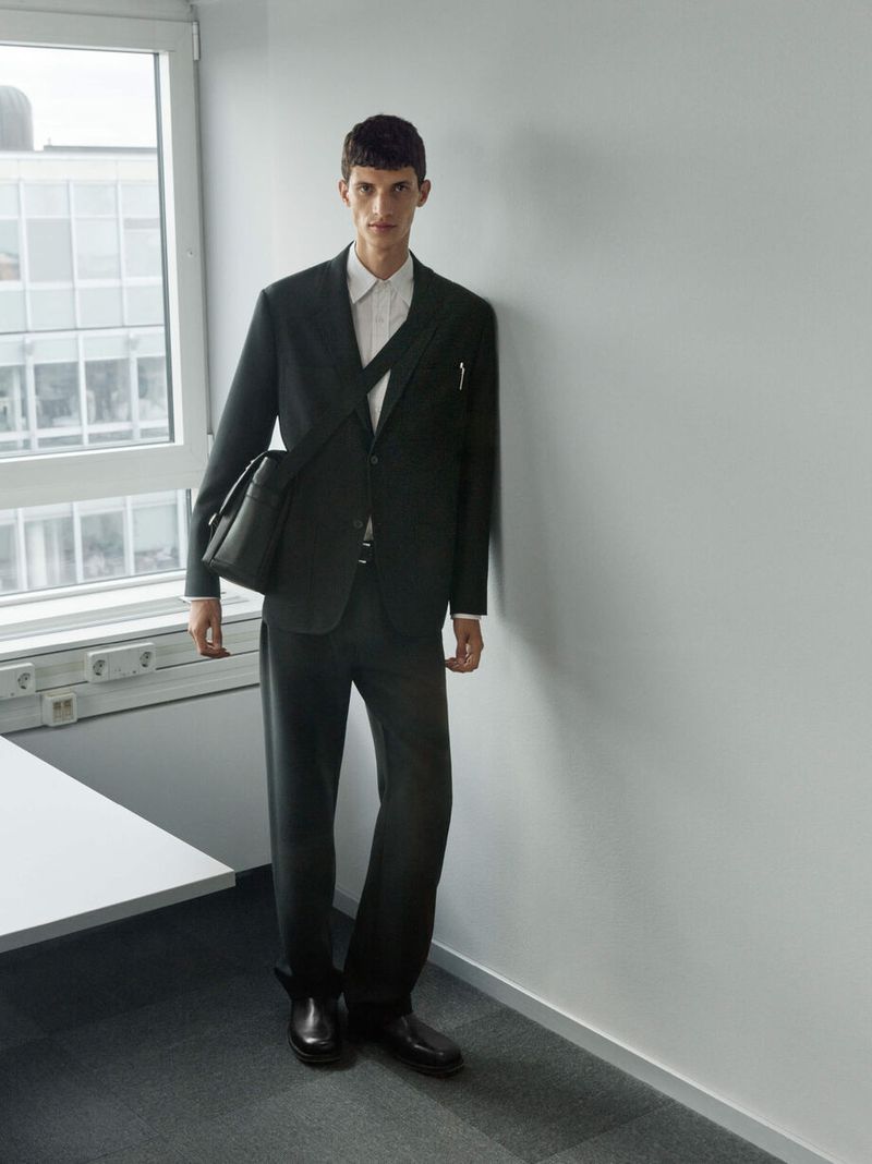 Stylish man in a dark suit, posing indoors with a shoulder bag. Professional attire, fashion-forward, modern office environment with natural light.