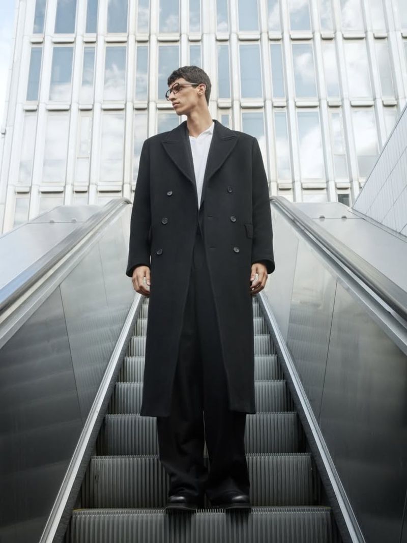 Man in black coat poses on escalator against a tall modern building. Wearing glasses and stylish outfit. Fashion editorial and urban scene.